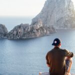 Man enjoys a scenic view of Es Vedrà at sunset from a cliff in San Juan Bautista, providing a perfect summer escape.
