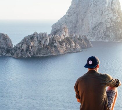 Man enjoys a scenic view of Es Vedrà at sunset from a cliff in San Juan Bautista, providing a perfect summer escape.